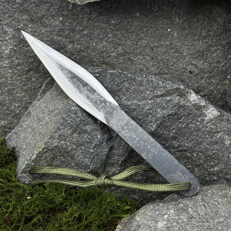 May include: A hand-forged, silver-colored throwing knife with a sharp, pointed blade. The handle is a flat, textured metal with a small hole at the end. A green cord is attached to the handle. The knife rests on a gray rock surface.
