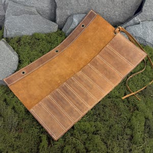 May include: Open, tan leather tool roll with multiple pockets and a leather tie closure. The roll is made of distressed leather and features white stitching. The tool roll is displayed on a bed of green moss.