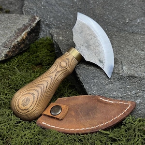 Op de afbeelding: Een mes met een gebogen lemmet, houten handvat en leren schede. Het lemmet is zilverkleurig, het handvat lichtbruin met een houtnerfpatroon en de schede bruin met witte stiksels en een drukknoopsluiting.