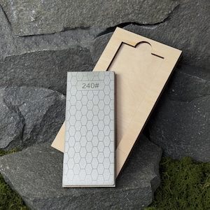 Puede incluir: Una piedra de afilar plateada con un patrón de panal y el número "240#" impreso. Descansa sobre una base de madera. El fondo presenta una superficie de piedra natural.