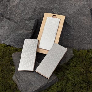 Pode incluir: Tr&ecirc;s pedras de amolar com um padr&atilde;o de favo de mel s&atilde;o exibidas em uma superf&iacute;cie rochosa. Uma pedra est&aacute; em uma moldura de madeira, e as outras est&atilde;o soltas. As pedras s&atilde;o rotuladas com os n&uacute;meros 240#, 600# e 1200#.