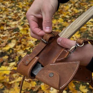 May include: A brown leather axe sheath with white stitching and a metal clasp. The sheath is designed to hold a small axe with a wooden handle. The axe head is visible inside the sheath.