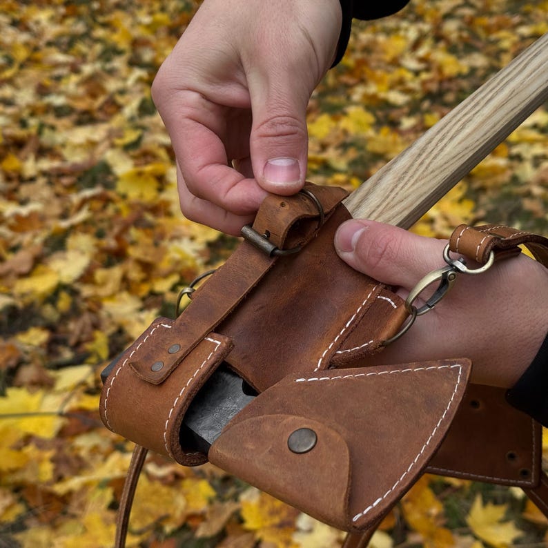 May include: A brown leather axe sheath with white stitching and a metal clasp. The sheath is designed to hold a small axe with a wooden handle. The axe head is visible inside the sheath.