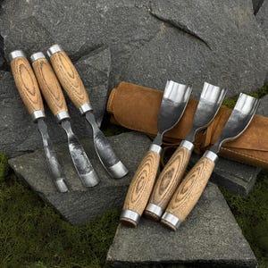 May include: A set of six wood carving gouges with curved steel blades and light brown wooden handles featuring visible grain patterns. The tools are arranged with three resting on dark gray stone and three placed on a brown leather tool roll.