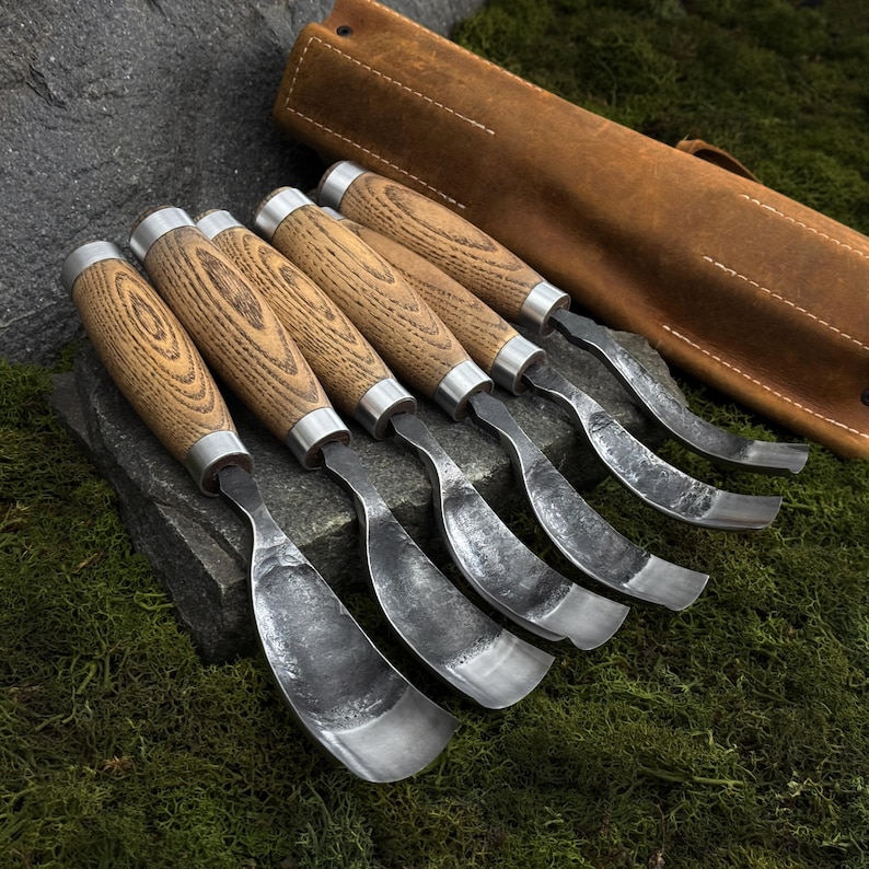 May include: A set of five wood carving gouges with curved, hammered metal blades and light brown wooden handles with visible grain patterns. The tools feature silver-colored metal ferrules and are accompanied by a brown leather tool roll.