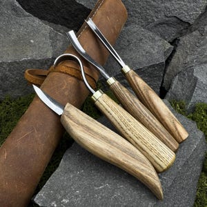 May include: A set of four wood carving tools with wooden handles and metal blades, displayed with a brown leather tool roll. The tools have various blade shapes, including a hook knife and a curved gouge. The wood grain varies in color from light to dark.