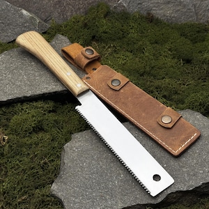 Może przedstawiać: Piła ręczna z jasnym drewnianym uchwytem i ostrzem ze stali nierdzewnej, spoczywająca na szarej kamiennej powierzchni. Piła jest wyposażona w brązową skórzaną pochwę z zatrzaskami. Ostrze piły ma około 30 cm długości.
