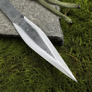 May include: Close-up of a hand-forged spearhead with a sharp, pointed tip. The metal blade has a textured, gray finish and a polished edge. A green and black rope is visible in the background, resting on a stone and green moss.