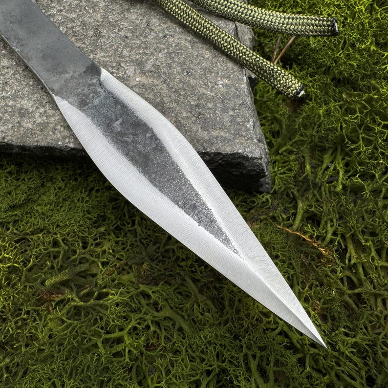 May include: Close-up of a hand-forged spearhead with a sharp, pointed tip. The metal blade has a textured, gray finish and a polished edge. A green and black rope is visible in the background, resting on a stone and green moss.