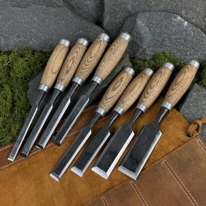 May include: A set of eight woodworking chisels with light brown wood handles and silver metal ferrules, featuring dark steel blades of varying widths. The chisels are arranged in a row on a brown leather tool roll with visible stitching.