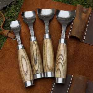 May include: Set of five wood carving tools with wooden handles. The tools have a curved blade and are arranged on a brown leather surface.