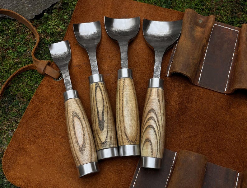 May include: Set of five wood carving tools with wooden handles. The tools have a curved blade and are arranged on a brown leather surface.