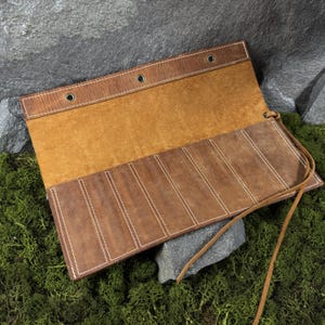 May include: Open leather tool roll in shades of brown, with white stitching and a tan interior. The roll has multiple pockets and a leather tie closure. The tool roll is displayed on a bed of green moss and rocks.