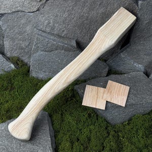 Könnte beinhalten: Ein hellfarbener Holzbeilgriff mit geschwungenem Design und zwei kleinen Holzquadraten. Der Griff ist glatt und scheint aus Hartholz gefertigt zu sein. Der Hintergrund zeigt graue Steine und grünes Moos.