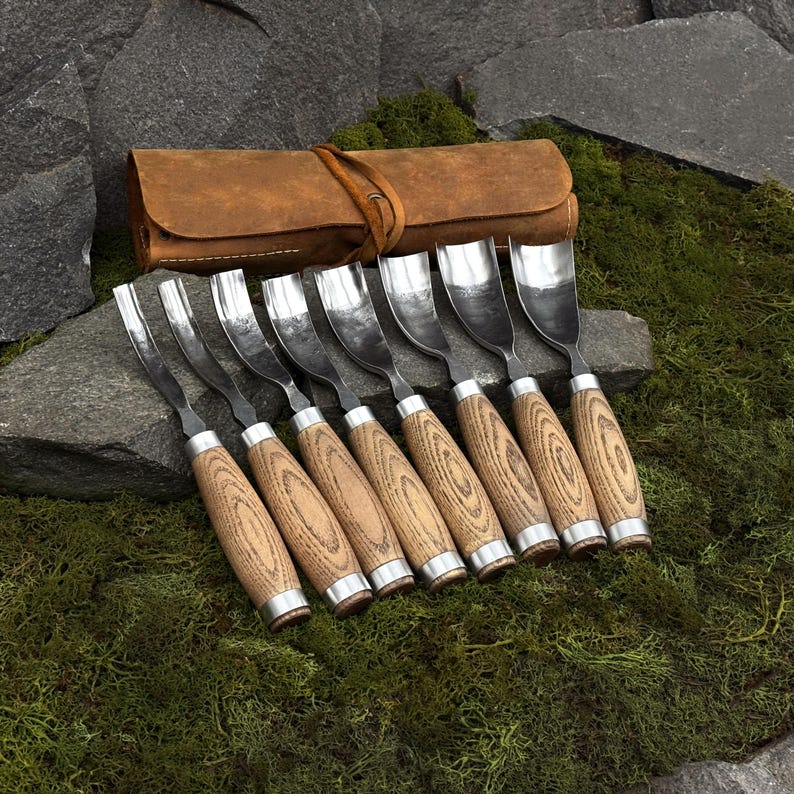 May include: A set of eight wood carving gouges with curved metal blades and light brown wooden handles, arranged in a row. The tools are accompanied by a brown leather roll-up case with a tie closure, featuring a visible grain texture.
