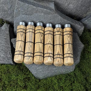 Könnte beinhalten: Sechs Holz-Stifthalter mit hellem Holzmaserungsmuster und schwarzen Bändern. Jeder Halter hat eine silberfarbene Metalloberseite. Die Halter sind auf einer grauen Steinoberfläche mit grünem Moos angeordnet.