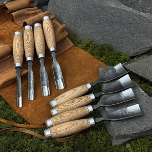 May include: A collection of wood carving chisels with wooden handles and metal blades. The chisels are displayed on a brown leather tool roll and a bed of green moss and gray rocks. The chisels are designed for woodworking.
