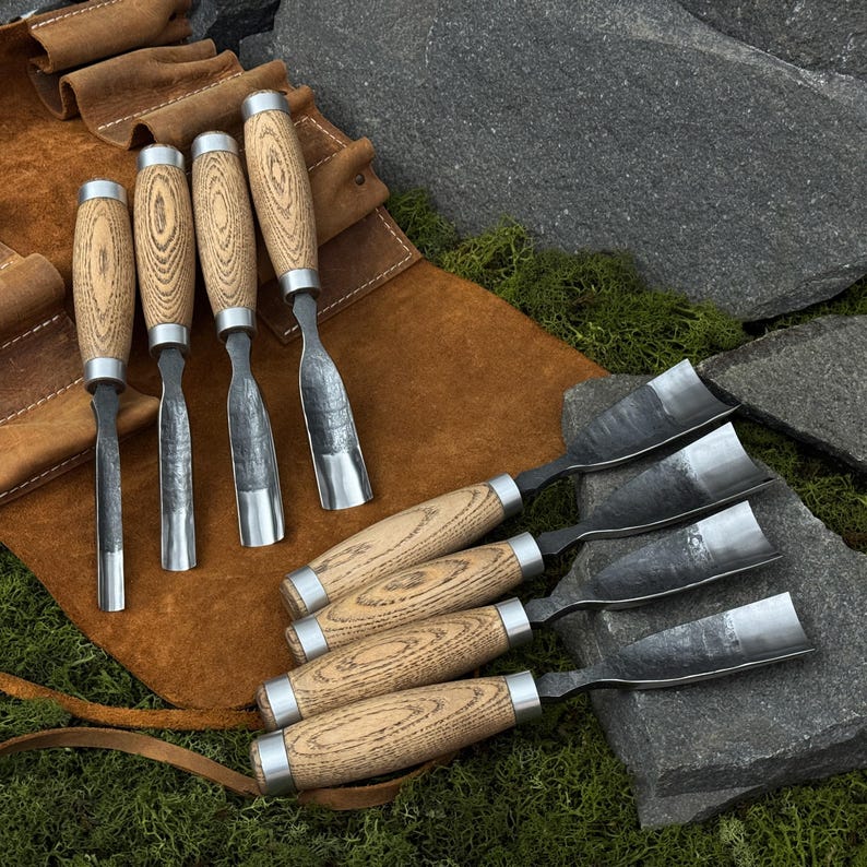 May include: A collection of wood carving chisels with wooden handles and metal blades. The chisels are displayed on a brown leather tool roll and a bed of green moss and gray rocks. The chisels are designed for woodworking.