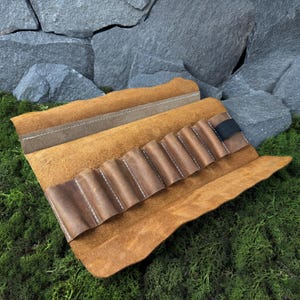 Peut inclure: Un étui à stylos en cuir marron ouvert, révélant une série de poches cousues. L'intérieur est d'une teinte plus foncée de marron. Une bande élastique noire maintient l'étui fermé. Le cuir a un aspect naturel et texturé.