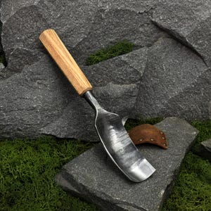 May include: A hand trowel featuring a dark, forged metal blade with a visible hammered texture and a smooth, light-toned wooden handle. A small, brown leather sheath with metal rivets is positioned near the base of the metal blade.