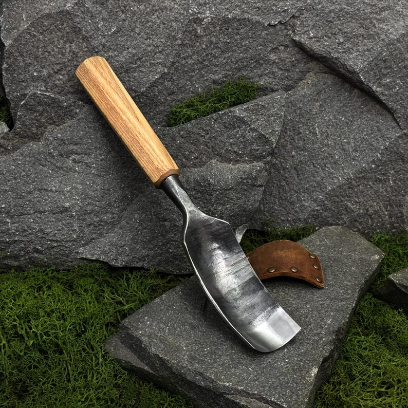 May include: A hand trowel featuring a dark, forged metal blade with a visible hammered texture and a smooth, light-toned wooden handle. A small, brown leather sheath with metal rivets is positioned near the base of the metal blade.