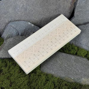 Peut inclure: Un objet rectangulaire en bois clair avec trois rangées de motifs géométriques. Chaque rangée présente un motif différent de triangles et de formes en étoile. L'objet repose sur de la mousse verte et des roches grises.