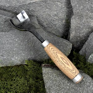 May include: A hand tool featuring a curved, polished metal blade with a dark, textured finish and a cylindrical wooden handle. The handle displays a prominent light brown wood grain pattern and is secured by two smooth, silver-toned metal ferrules.