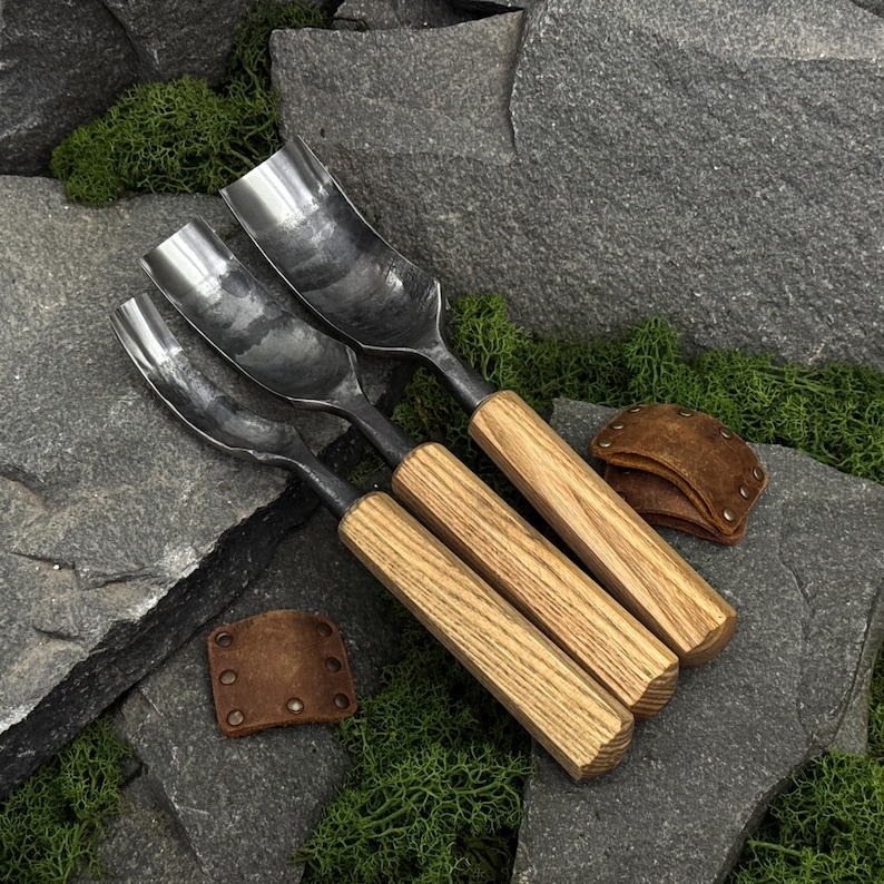 May include: Three metal gouges with curved, polished steel blades and octagonal wooden handles. Each tool features a distinct width, and two small, brown leather blade guards with metal rivets are positioned near the handles on the stone surface.