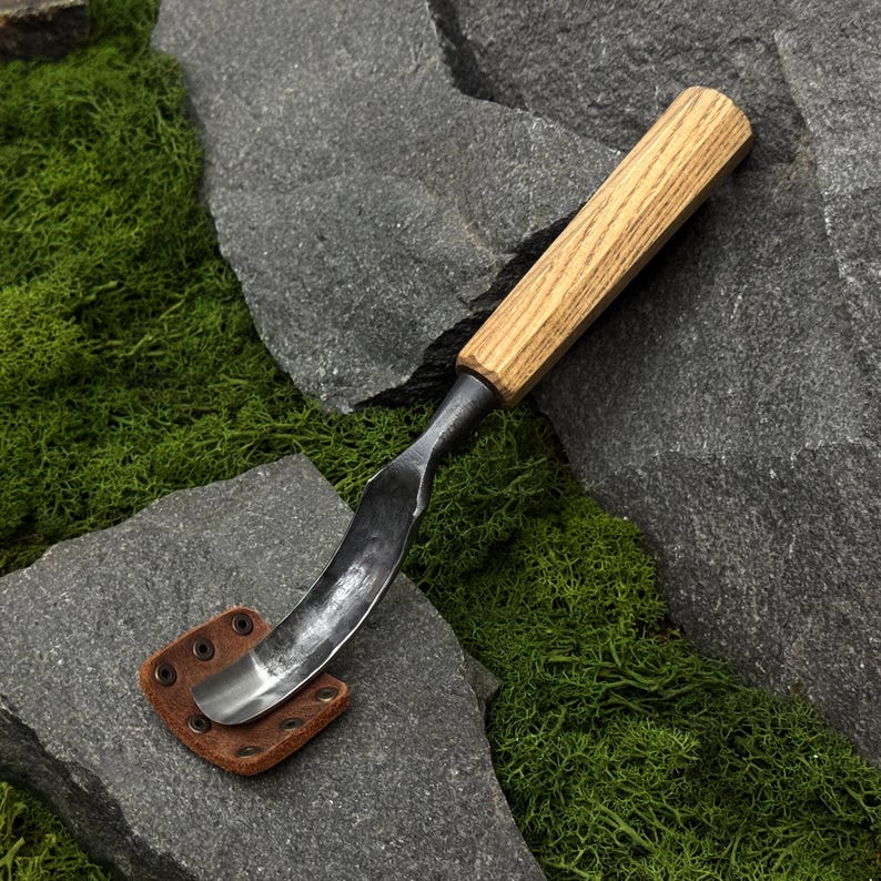 May include: A wood carving tool featuring a curved, polished metal blade and a light-colored wooden handle. The blade is protected by a small, square brown leather sheath secured with four metal rivets, showcasing a functional design for woodworking.