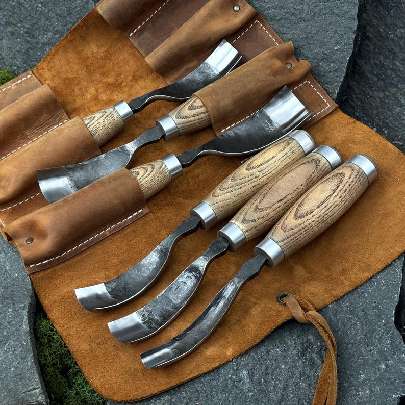 May include: A set of five metal woodworking gouges with curved blades and light-colored wooden handles, arranged in a brown leather tool roll. Each tool features a polished metal ferrule connecting the blade to the textured wood handle.