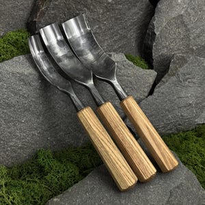 May include: Three metal gouges with curved, polished steel blades and light brown wooden handles. Each tool features a distinct width, showcasing a metallic finish with visible texture on the steel, arranged side by side in a linear formation.