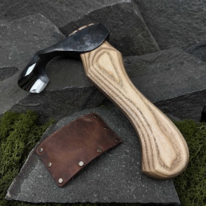May include: A hand-forged wood carving hook with a brown leather sheath. The hook has a curved blade and a wooden handle with a light wood grain.