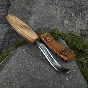 May include: A wood-handled carving knife with a curved blade, resting on a gray stone surface. The knife is accompanied by a brown leather sheath with two brass-colored button closures. The handle has a natural wood grain.