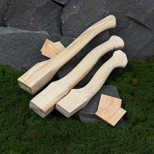 May include: Three unfinished wooden axe handles with a natural wood grain. Two small wooden squares are also present. The handles are arranged diagonally, with the largest at the top. The background features dark grey rocks and green moss.