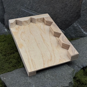 Könnte beinhalten: Ein helles Holztablett mit geometrischem Randdesign. Das Tablett ist rechteckig mit natürlicher Holzmaserung. Der Rand weist eine Reihe von winkligen Aussparungen auf. Das Tablett liegt auf einer Steinoberfläche.