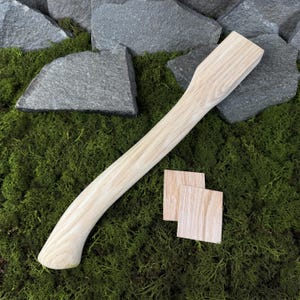 Puede incluir: Un mango de hacha de madera clara con un agarre curvo y una cabeza rectangular. También hay dos pequeños trozos de madera cuadrados. Los artículos descansan sobre musgo verde y están rodeados de piedras grises.