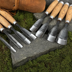May include: A collection of wood carving chisels with wooden handles and metal blades. The chisels are arranged on a gray stone surface with green moss in the background. A leather pouch is visible in the upper left corner.