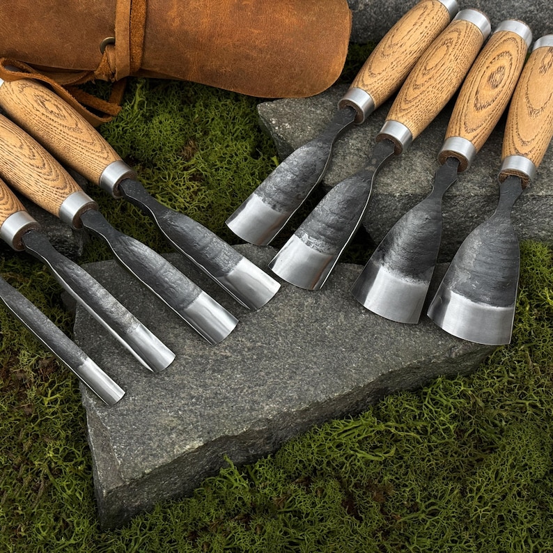 May include: A collection of wood carving chisels with wooden handles and metal blades. The chisels are arranged on a gray stone surface with green moss in the background. A leather pouch is visible in the upper left corner.