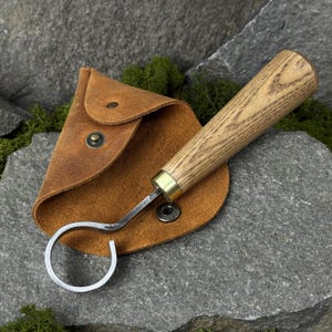 May include: A wood-handled leather tool with a curved metal end, resting on a brown leather pouch. The tool has a brass ferrule and a light-colored wooden handle. The pouch is secured with a snap closure. The background is a gray stone.
