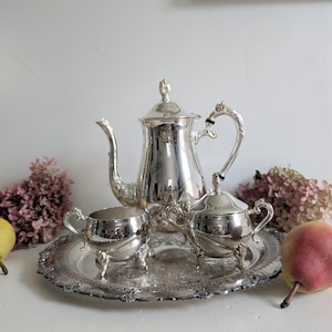 May include: A silver-plated tea service set comprising a tall teapot, a creamer, and a lidded sugar bowl, all featuring ornate handles and feet. The three pieces are arranged on a matching round silver-plated serving tray with an embossed rim.