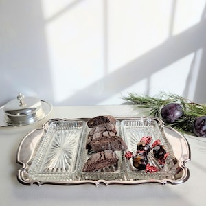May include: A silver serving tray with three glass compartments, holding biscotti and sweets. A silver butter dish is on the left. Plums and greenery suggest a festive theme.