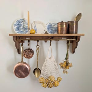 May include: A wooden kitchen shelf with various items. Blue and white plates, copper pots, and a net bag of lemons are visible. A candle and dried flowers add to the rustic aesthetic. The shelf is a charming display.