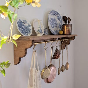 May include: A wooden shelf with a brass rod and hooks, displaying blue and white plates, a vase, and copper kitchenware. Below, various kitchen utensils and a net bag hang from the hooks, creating a vintage kitchen aesthetic.