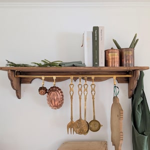 Peut inclure: Une étagère en bois avec une barre en laiton où sont suspendus des ustensiles de cuisine. Des moules en cuivre et des outils de cuisine en laiton sont suspendus en dessous. Livres, récipients en cuivre et bougies sur l'étagère. Une planche à découper en bois et un tablier vert sont également visibles.