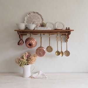 May include: A wooden wall shelf with a brass rail holding copper molds, a copper pan, a brass pan, and three brass ladles. The shelf holds two white bowls, a silver tray, two white plates, and small shakers. A white pitcher with dried flowers sits below.