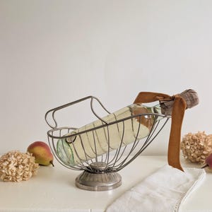May include: A silver-toned metal wire wine cradle holding a clear glass bottle. The bottle neck is wrapped in a brown velvet ribbon. The cradle features a curved handle and a circular base, designed to hold a bottle at an angled position.