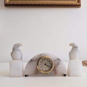 May include: A vintage marble clock with two eagle sculptures. The clock has a gold-coloured face with black numbers and hands. The eagles are white with grey markings, and the clock base is a light pink and white marble.
