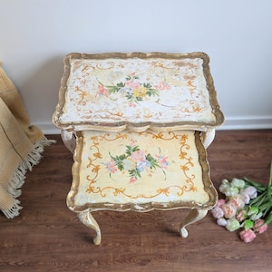 May include: Two-piece nesting table set with a distressed, antique aesthetic. The tables feature a cream-colored base with floral designs in pink, yellow, and blue. Gold trim outlines the scalloped edges. The legs are curved and painted cream.