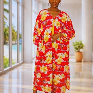 May include: A long, flowing red dress with a vibrant floral pattern of yellow flowers. The dress has long sleeves and a V-neckline. The model is wearing white high-heeled sandals. The dress is suitable for a beach cover-up or resort wear.