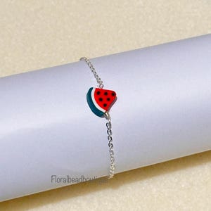 Può includere: Un braccialetto a catena d'argento con un ciondolo a forma di anguria. Il ciondolo è rosso, verde e bianco con semi neri.